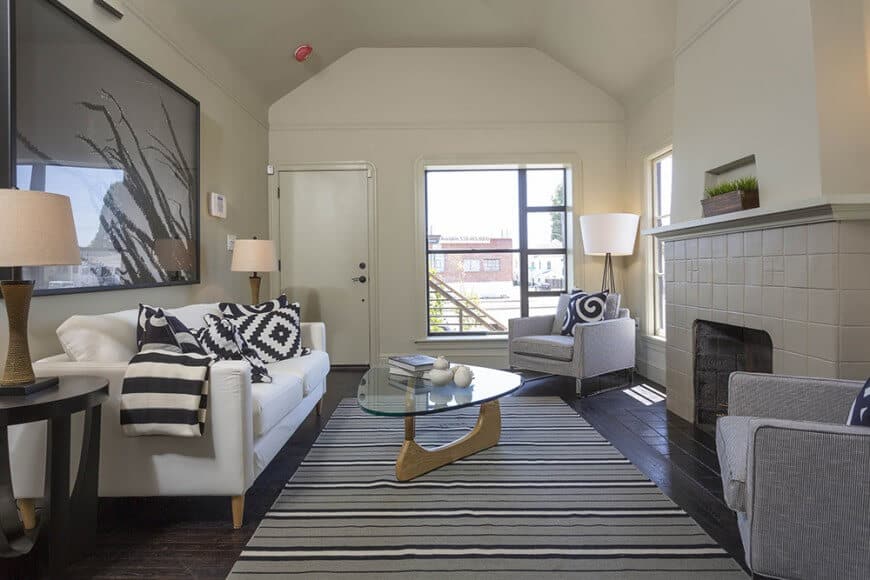 A glass top coffee table sits on the striped area rug flanked by a tiled fireplace and white sofa that's accented with patterned pillows. It is accompanied by gray armchairs and round side tables topped with traditional lamps.