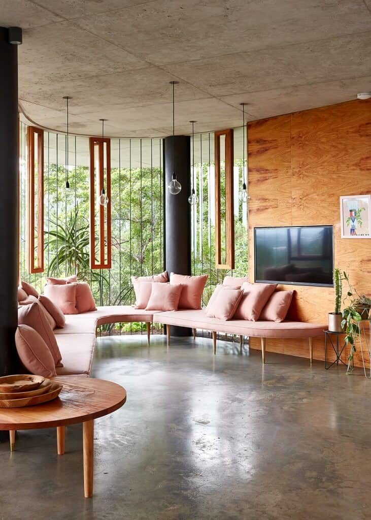 Airy living room with a curved sofa and fireplace fitted on the wood paneled wall. It has concrete flooring and a distressed ceiling mounted with bulb pendant lights.