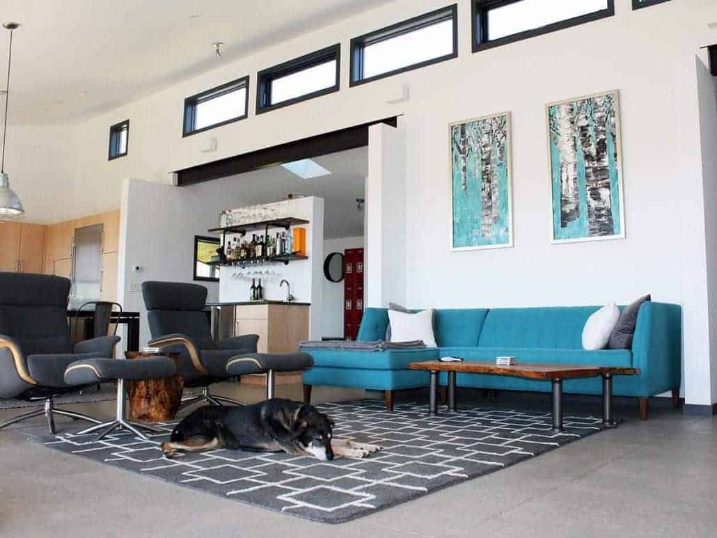Blue artworks and sofa stand out against the white walls that are fitted with aluminum-framed windows. This room showcases black lounge chairs and a patterned rug that lays on the tiled flooring.