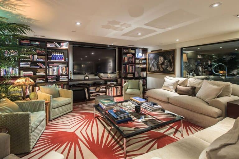 A red patterned rug stands out in this living room with cozy seats and a glass top coffee table facing the wall mount TV surrounded by open shelving.