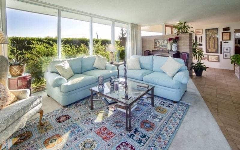 Medium-sized living area with a gray wingback seat and blue couches paired with a glass top coffee table over a printed rug. It has carpet flooring and a panoramic window overlooking the outdoor greenery.