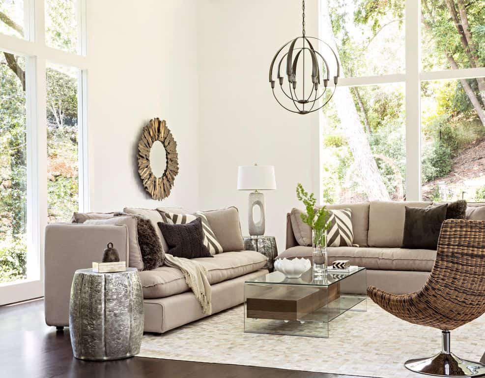 Gray sofas and a wicker chair surround a glass coffee table lighted by a spherical pendant. This room is decorated with a sunburst mirror and a stylish table lamp that sits on a barrel side table.