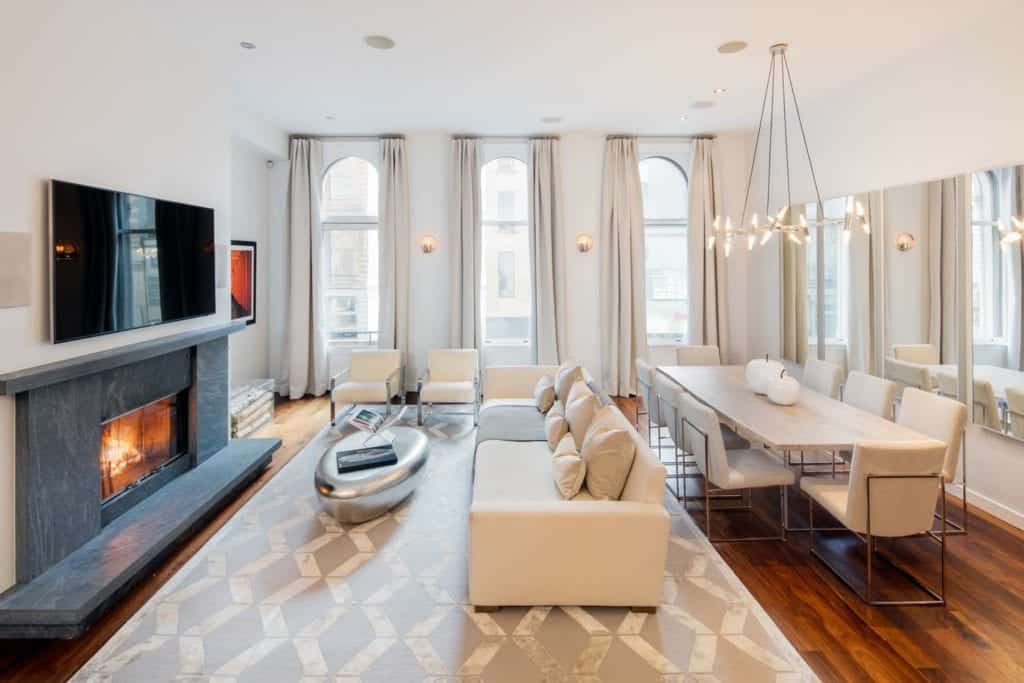 The modern living room offers a fireplace with a wall-mount TV on top along with white seats paired with a chrome coffee table over a patterned area rug. It has rich hardwood flooring and arched windows covered in gray draperies.
