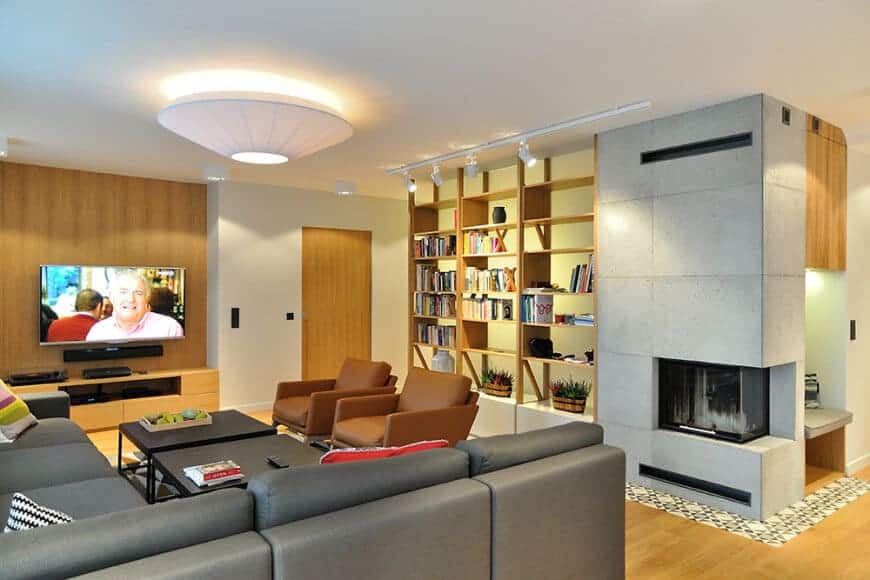 Gray sectional sofa and brown leather armchairs surround a pair of black coffee tables facing the wall mount TV that hung above the wooden console table. It is accompanied with open shelving and a modern fireplace fitted on the concrete paneled pillar.