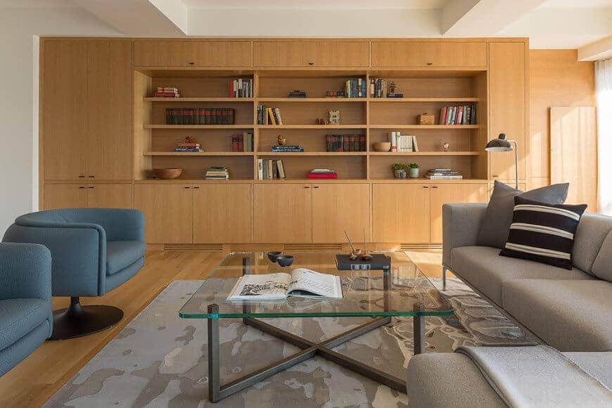 The cozy living room offers sleek seats and a glass top coffee table over a printed area rug. It includes built-in cabinets with open shelving filled with books and decors.