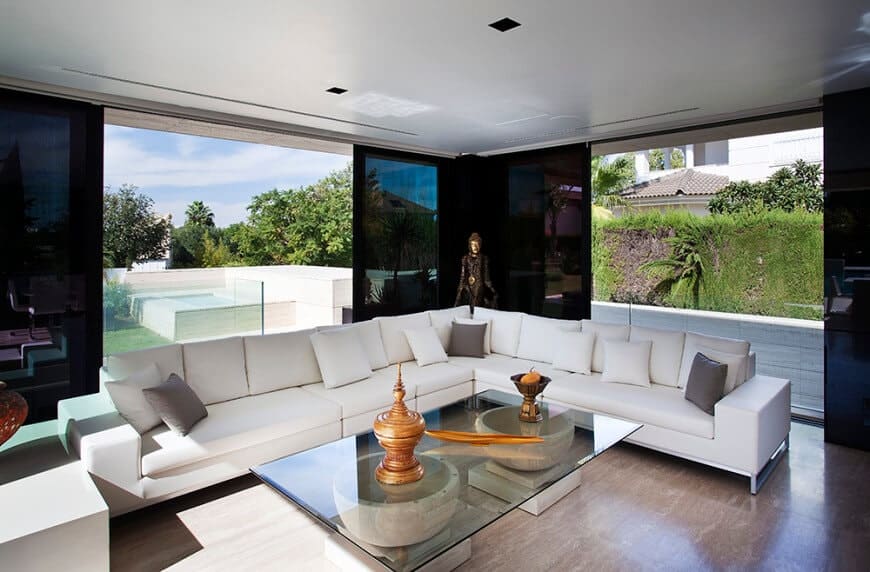 Modern living room with a glass top coffee table and a V-shaped sectional sofa filled with white and gray pillows. It has hardwood flooring and glass sliders leading out to the yard.