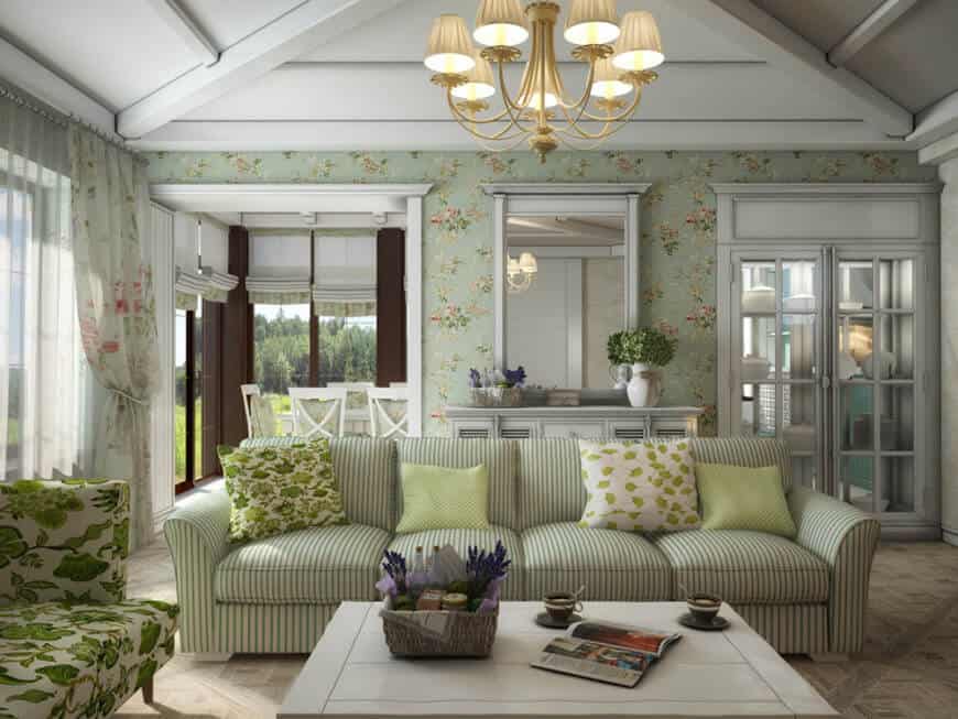 Clad in floral wallpaper, this living room showcases a striped sectional and green floral seat paired with a white coffee table. It includes brass chandelier and a framed mirror mounted above the louvered console table.