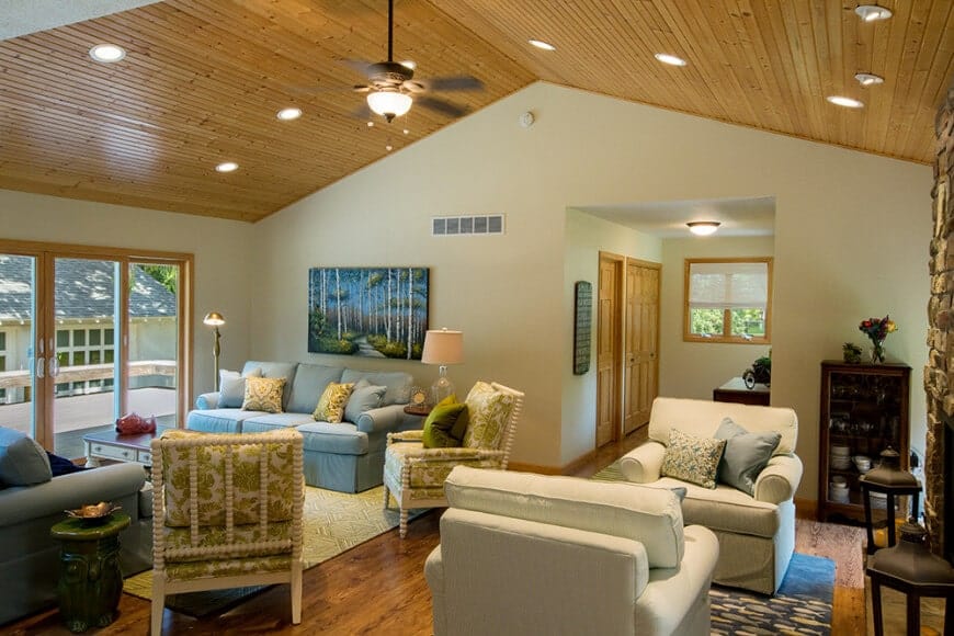 This living room features multiple seating areas and a fireplace fixed on the stone brick pillar. It is illuminated by a flush and recessed lights fitted on the cathedral ceiling clad in wood planks.