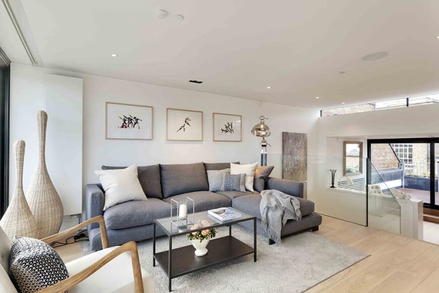 Minimalist wall arts hang above the L-shaped sectional accompanied by white armchair and a metal coffee table over a gray area rug. It has wide plank flooring and a regular ceiling mounted with recessed lights.