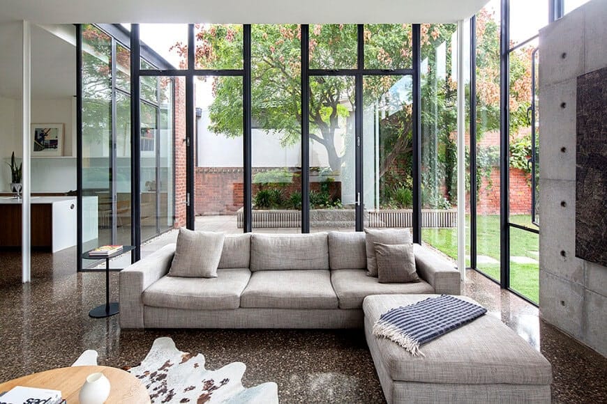 Plenty of natural light flows in through the full height glazing in this living room with a gray sectional sofa and a light wood coffee table that sits on a cowhide rug over the granite flooring.